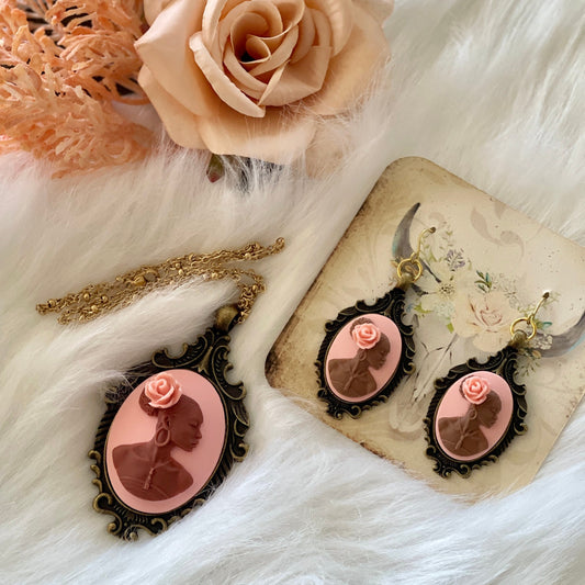 May include: A set of pink cameo jewelry with a gold chain necklace and earrings. The necklace and earrings feature a cameo of a woman&#39;s profile with a pink rose on her head. The cameo is set in a gold-toned oval frame.